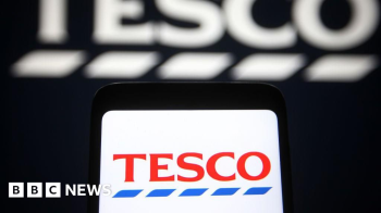 Getty Images O logotipo da Tesco  exibido em um smartphone, contra um fundo preto exibindo uma verso maior e desfocada do logotipo do supermercado.