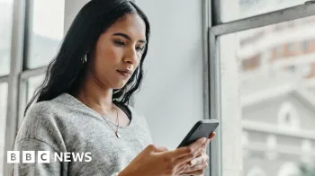 Getty Images Young woman in grey pullover standing by a window and looking at her phone with a serious look on her face.
