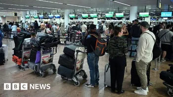 Filas de pessoas com carrinhos cheios de bagagens esperam em um terminal de aeroporto movimentado