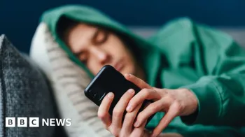 Um adolescente vestindo um moletom verde com o capuz levantado, deitado em almofadas em um sof� olhando para seu telefone celular