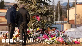 AFP via Getty Images Dois homens em p� do lado de fora, de costas para a c�mera, olhando para uma grande cole��o de flores e presentes deixados para as v�timas do tiroteio na escola, colocados embaixo de uma grande �rvore.