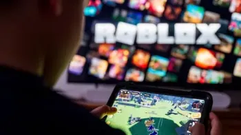Getty Images Uma vis�o por cima do ombro de uma crian�a segurando um tablet jogando no Roblox. Ao fundo, uma tela desfocada com o logotipo do Roblox.