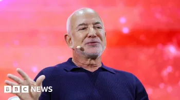 Jeff Bezos usando fone de ouvido com microfone e camiseta polo azul-marinho. Ele est� no palco em frente a um fundo vermelho brilhante