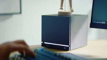 Steam Machine sobre uma mesa de madeira. Algu�m digita em um teclado, na frente de um monitor, ao lado dela.