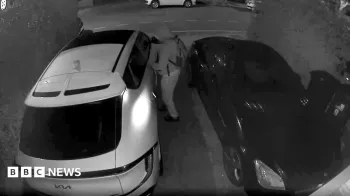 Um homem encapuzado parece estar mexendo em um carro Kia branco estacionado em frente a uma casa; a imagem foi capturada por uma c�mera de campainha.