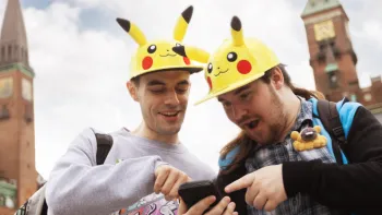 Dois f�s de Pok�mon Go usando chap�us em forma de Pikachu apontam e olham para um telefone, em choque.