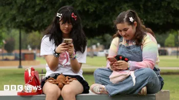 Duas jovens sentadas em um banco de cimento ao ar livre, cada uma olhando para o pr�prio celular.